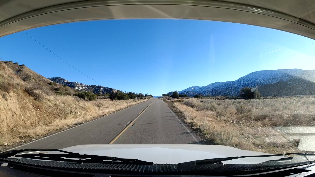 Dash-style view along a straight road in Coast California, with the vehicle hood at the bottom, dry grasses and scrub on both sides, mountains ahead, and no visible people.