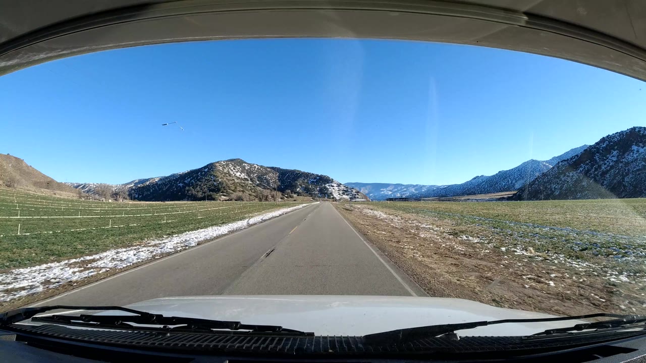 Forward vehicle view on a paved road in Coast California, passing green fields, roadside snow patches, brown hills, distant mountains, and no visible people.