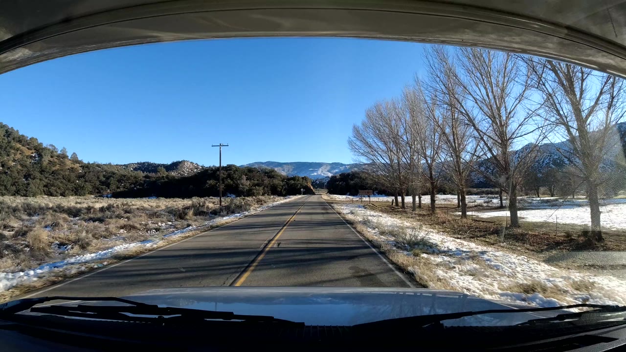 A Coast California two-lane road runs straight ahead past bare trees, utility poles, roadside snow, low brush, and distant hills, with no visible people.