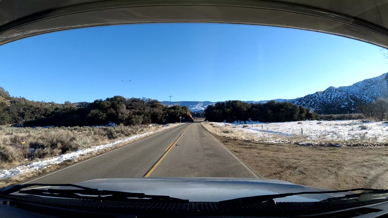The vehicle view continues along a Coast California road bordered by snow patches, scrubby vegetation, low hills, and a broad blue sky, with no visible people.