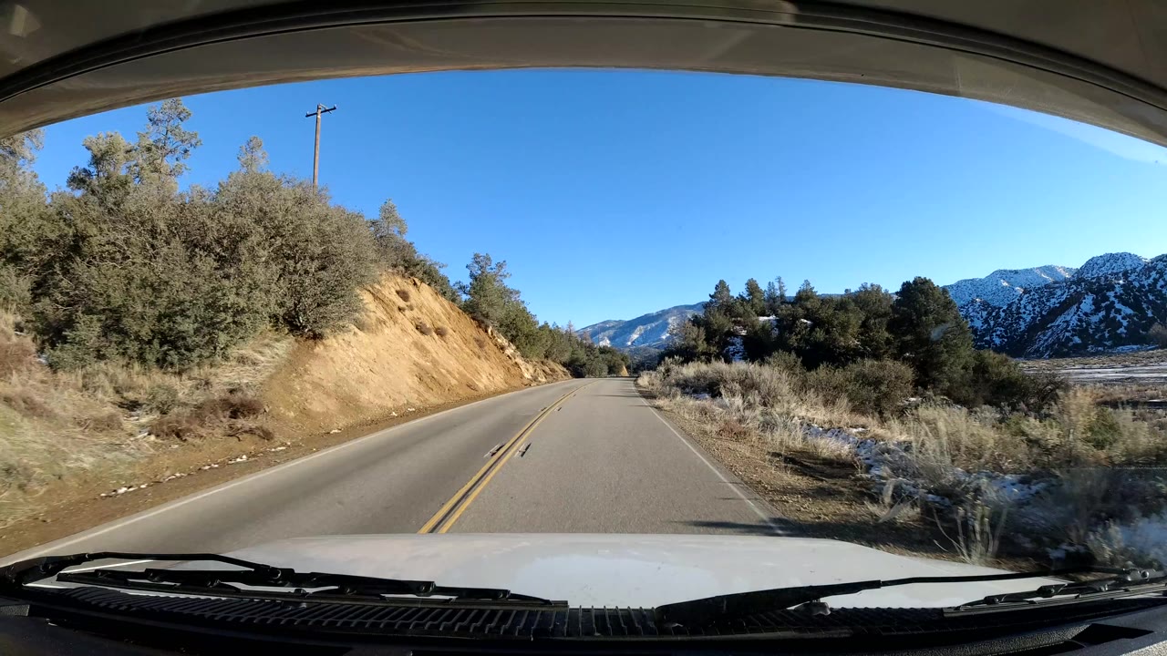 A Coast California road passes a tan roadside embankment and brush, with snow-dusted mountain slopes beyond the pavement and no visible people.