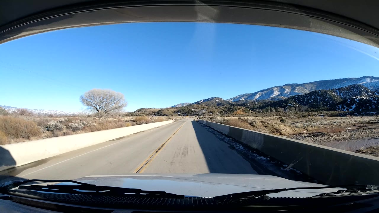 The vehicle looks down a Coast California road section with concrete side barriers, roadside scrub, snow patches, distant hills, and no visible people.