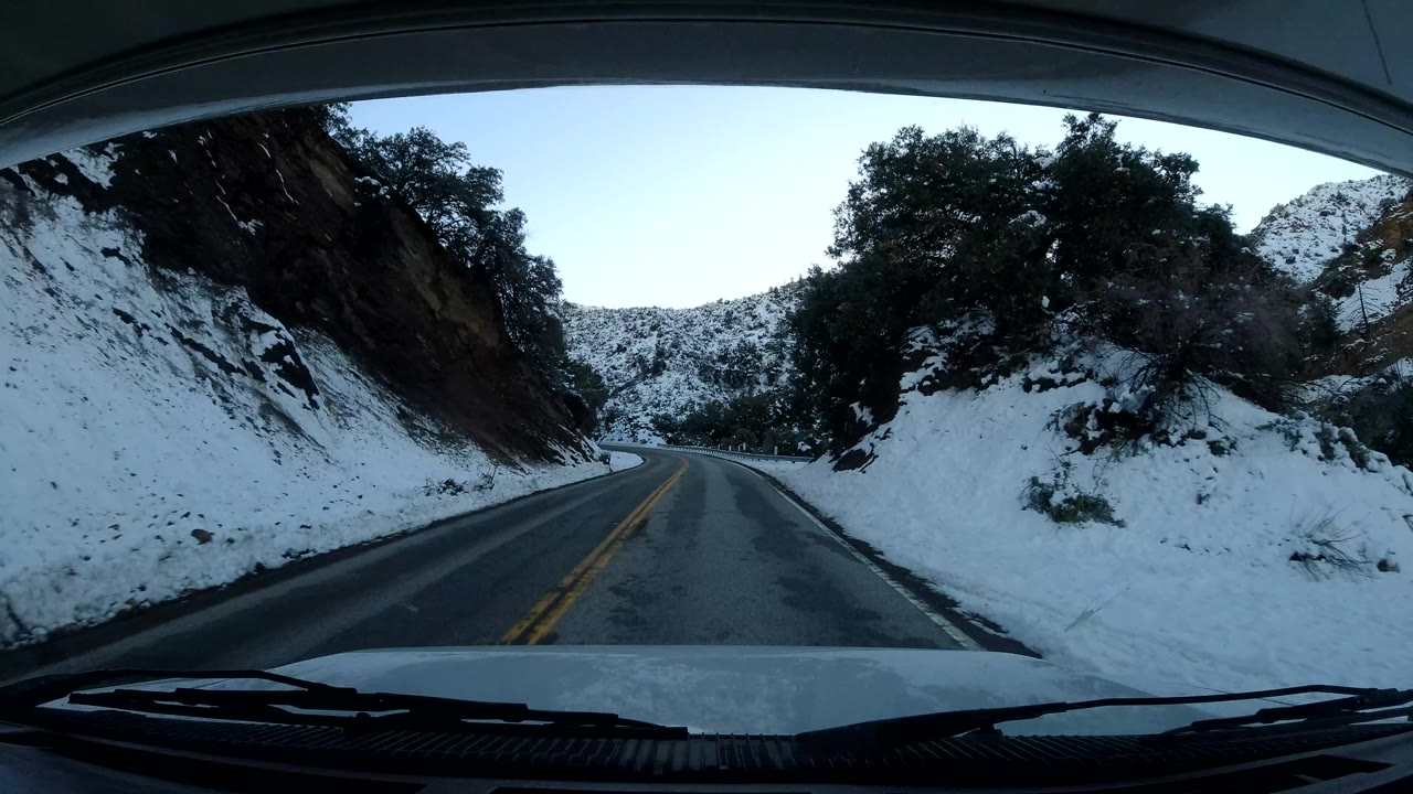 A Central Coast California road is seen through a vehicle windshield as it bends past snow-covered shoulders, dark trees, and a shaded hillside, with no visible people.