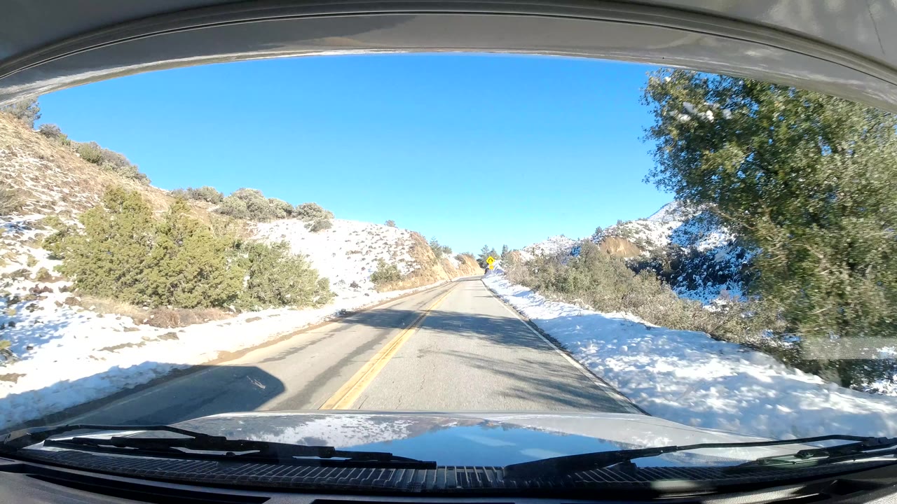 A brighter stretch of Central Coast California road shows snow on both shoulders, blue sky overhead, low shrubs and trees along the road, and no visible people.