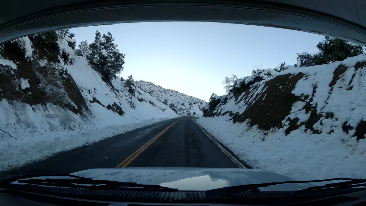 The vehicle viewpoint looks down a wet-looking two-lane road bordered by snow, with a sloped hillside on one side, distant tree-covered ridges, and no visible people.