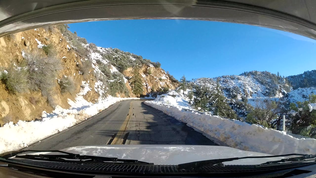 Coast California 2019 road view with the vehicle hood at the bottom, snow piled along both sides, a sunlit rock face to the left, distant hills, and no visible people.