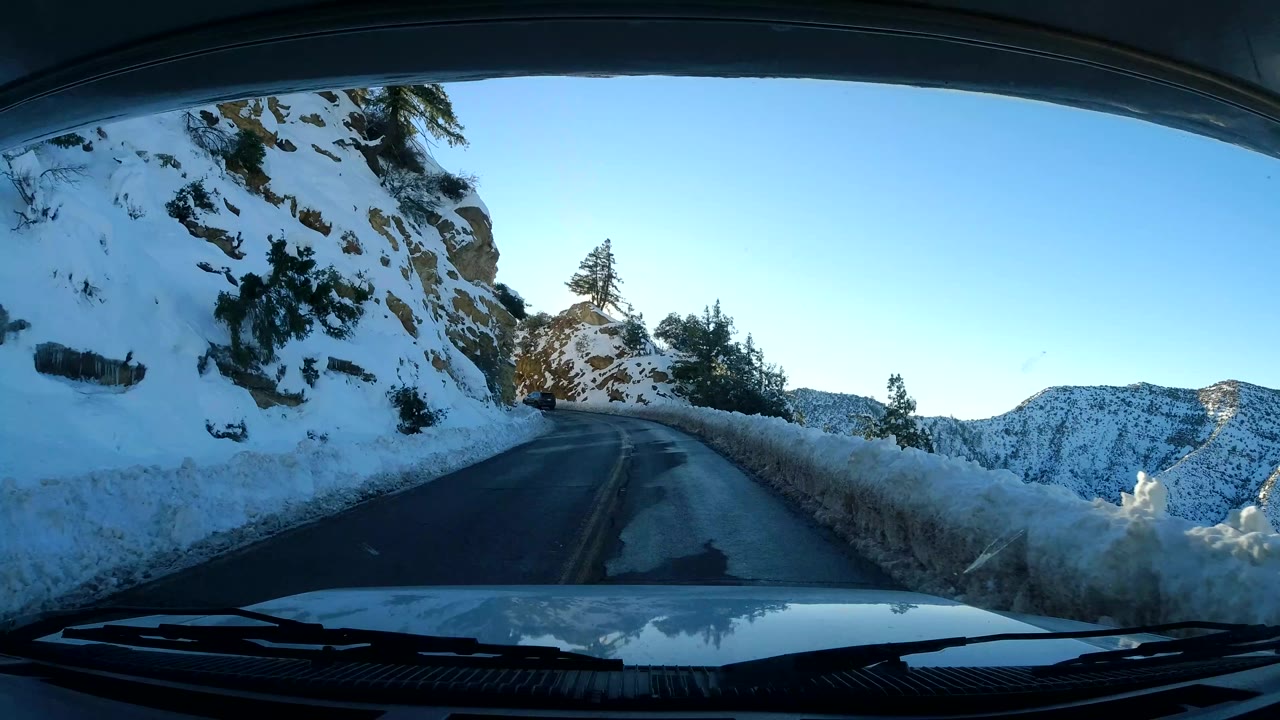 Coast California 2019 vehicle view of a left-bending road beside a snow-covered cut bank, with open snowy hills beyond the right edge and no visible people.