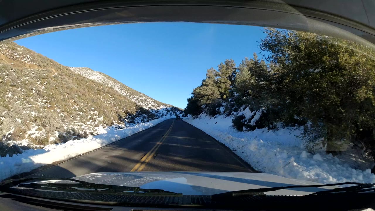 Central Coast California two-lane road seen from a vehicle, with snow piled along both shoulders, trees on the right hillside, blue sky above, and no visible people.