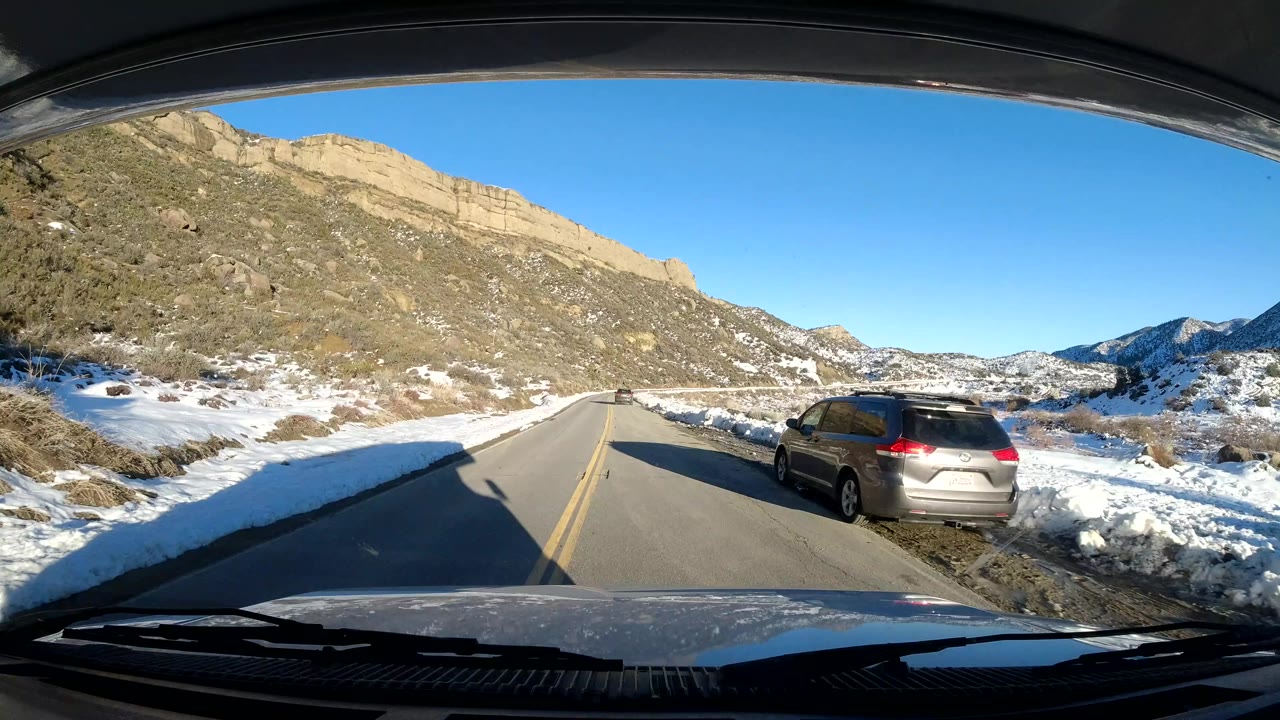 Central Coast California winter road scene with snow-covered shoulders, tan rocky hills, a gray SUV passing in the opposite lane, and no visible people.