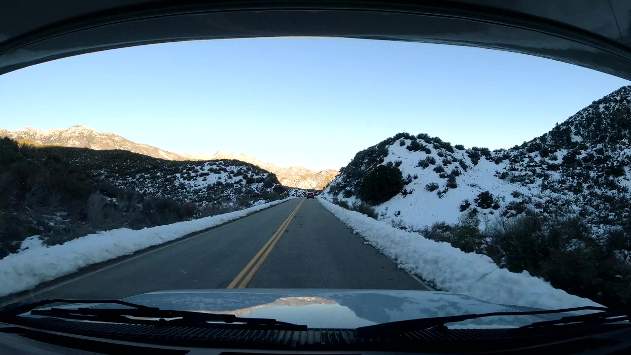 Windshield view in Coast California, United States, showing a straight two-lane road between snowbanks, dark hillsides, distant sunlit ridges, and no visible people.