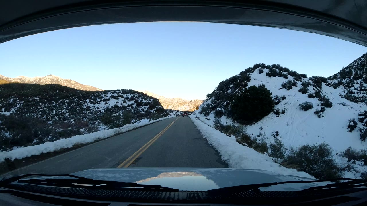 Windshield view in Coast California, United States, with the road running forward toward a slight bend, snow-covered shoulders, shrub-covered hills, and no visible people.