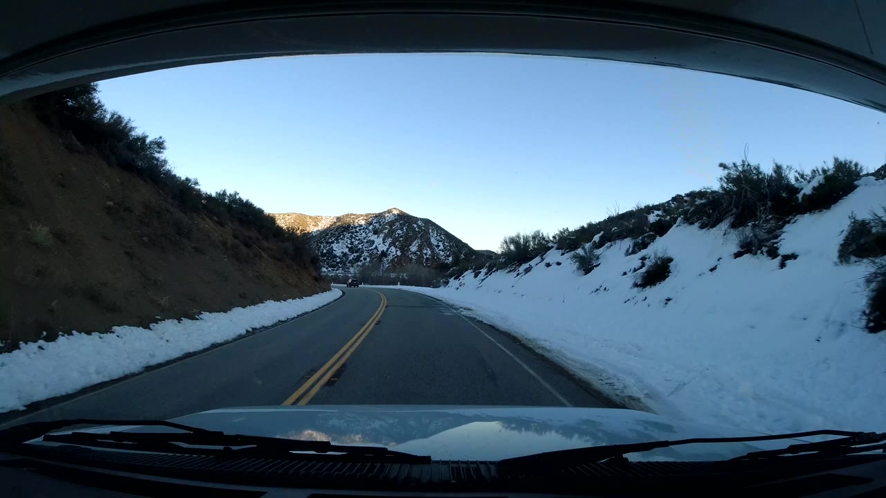 Windshield view in Coast California, United States, showing a road curving left between a dirt cut bank, snow-covered roadside brush, distant mountains, and no visible people.