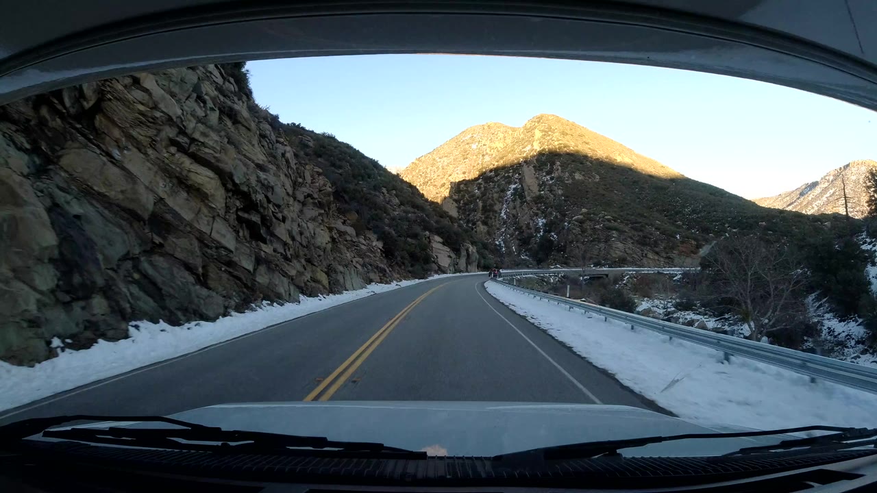 Vehicle-front view of a two-lane road in Coast California, United States, with snow on both shoulders, rocky slopes close to the road, sunlit hills ahead, and no visible people.