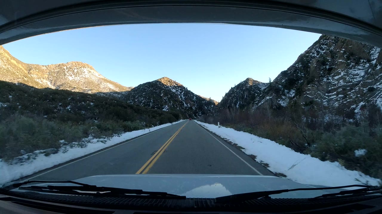 The vehicle view looks down an empty two-lane road between snow-covered brush and dark hills, with higher mountains catching light in the distance and no visible people.