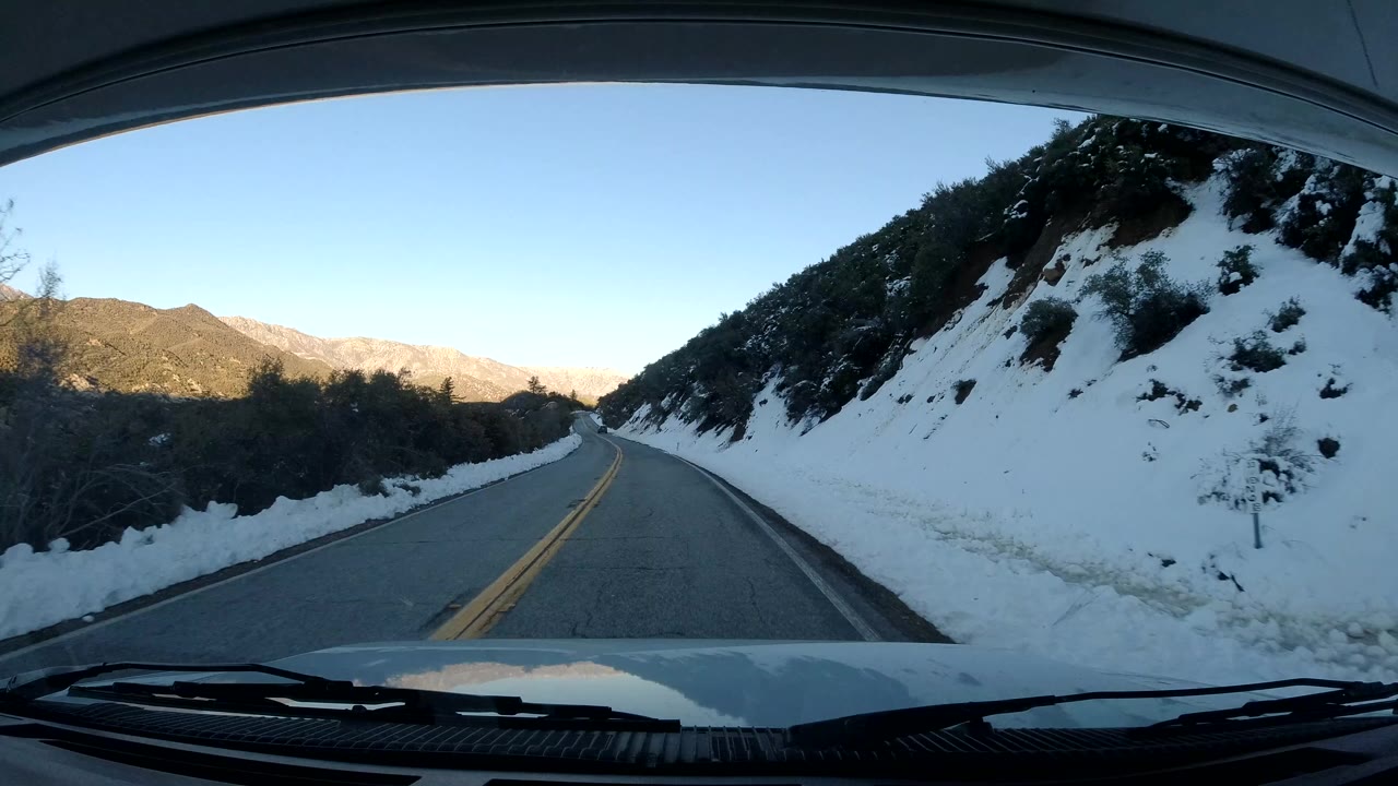 Vehicle POV on a Coast California two-lane mountain road, with snow piled along both shoulders, scrubby slopes ahead, the car hood at the bottom edge, and no visible people.
