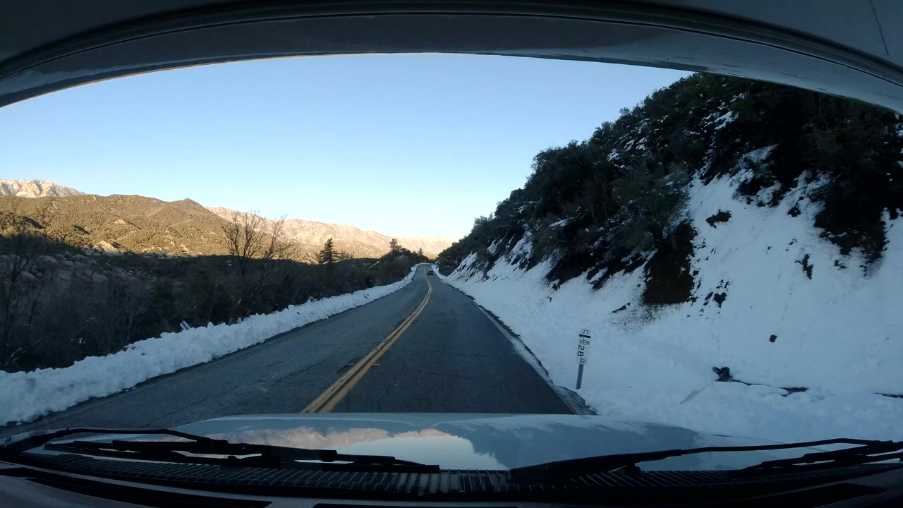 Vehicle POV in Coast California as the road rises beside a snow-covered hillside on the right, with distant mountains lit across the valley and no visible people.