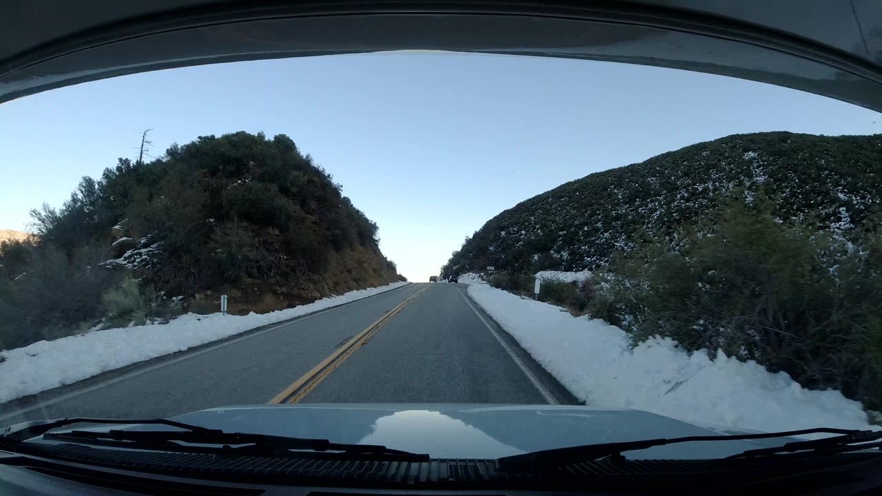 Vehicle POV on a Coast California road approaching a gentle curve, with brush-covered hills, patchy snow on both sides, clear blue sky, and no visible people.