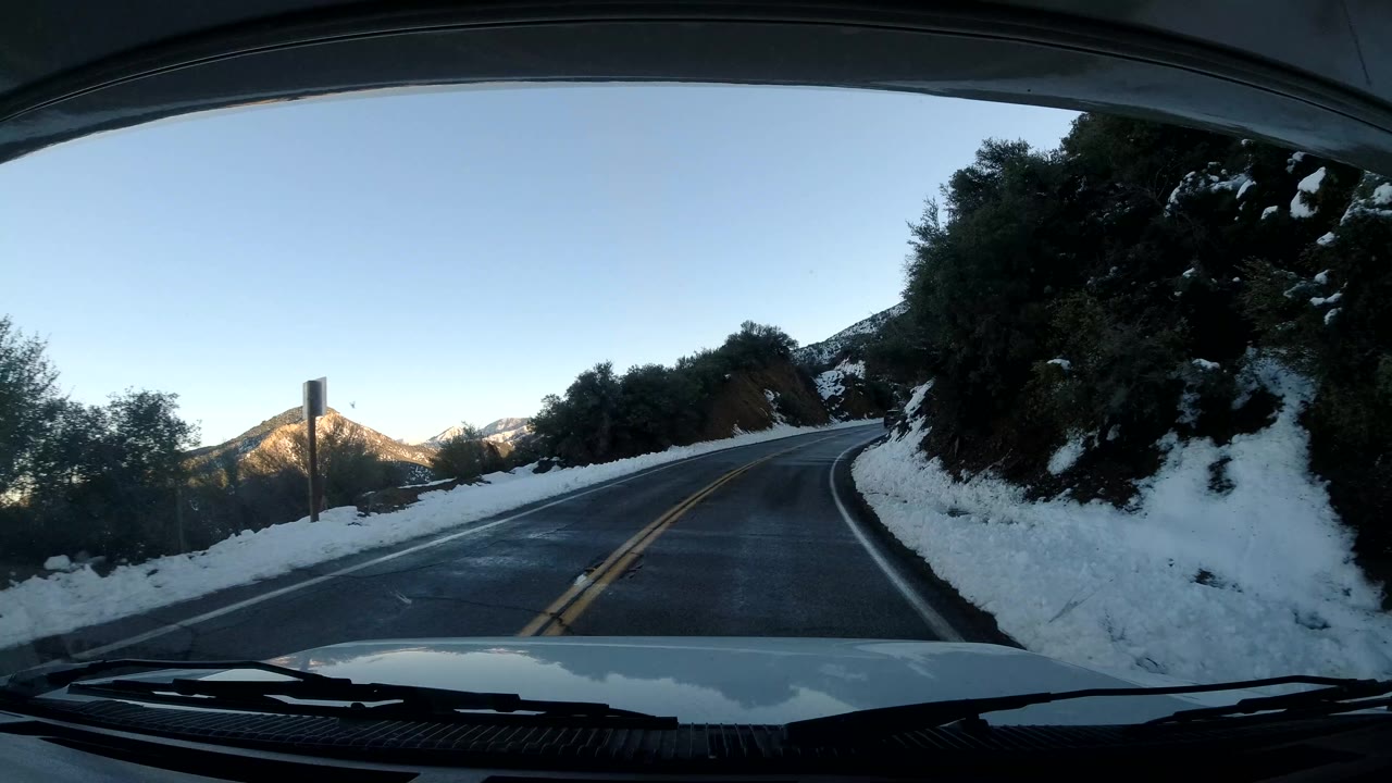 Vehicle POV in Coast California with the two-lane road bending left, dark brush and snowbanks close to the shoulders, mountains in the distance, and no visible people.