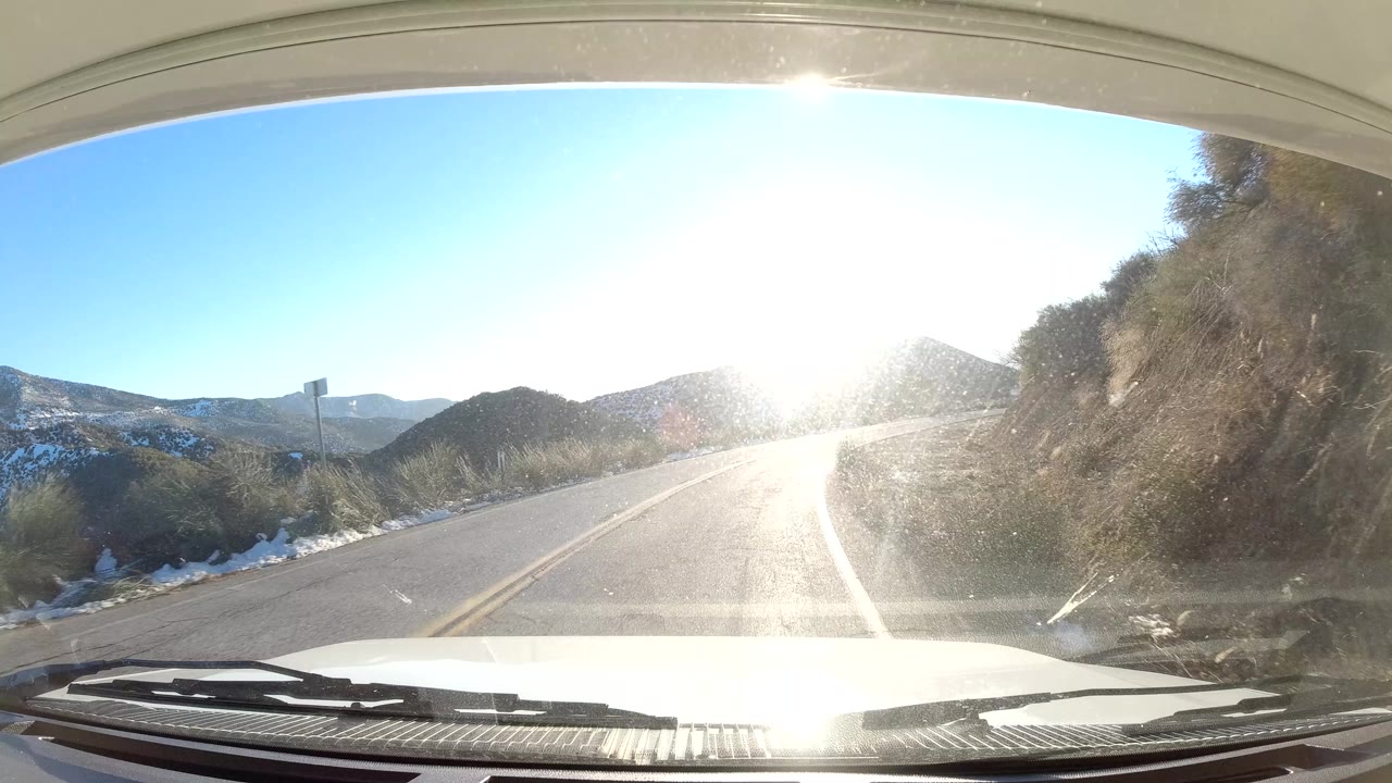 Forward windshield view on the California coast shows a two-lane road bending through dry hills with intense sun glare ahead and no visible people.
