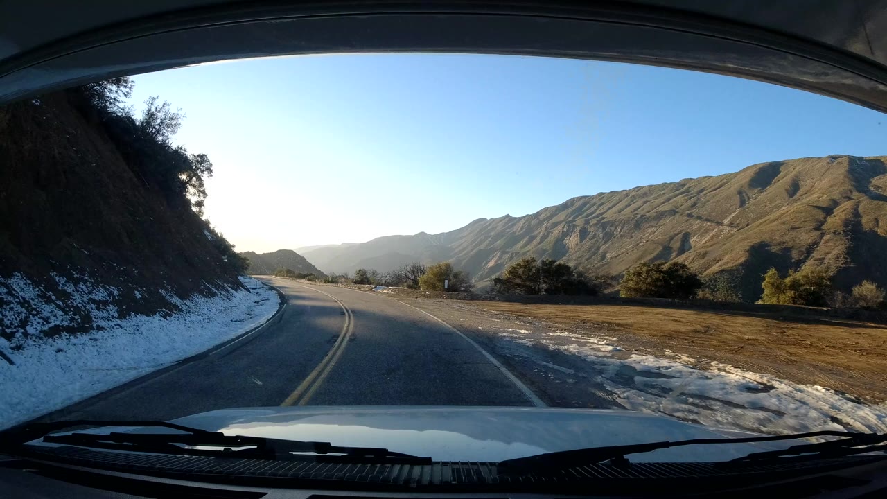A windshield view on the California coast shows the road curving past a dark hillside, snow along the shoulder, open valley terrain, and no visible people.