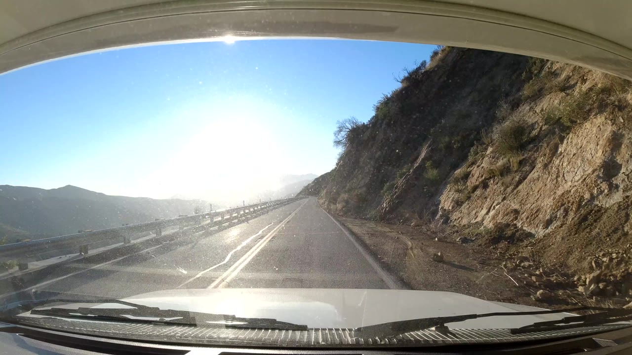 Two-lane road beside a rocky hillside with sun glare through the windshield.