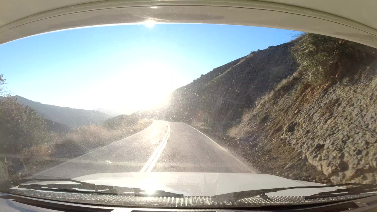 Windshield-level view in Coast California shows a two-lane mountain road running toward harsh low sun, with the vehicle hood and wipers at the bottom and no visible people.
