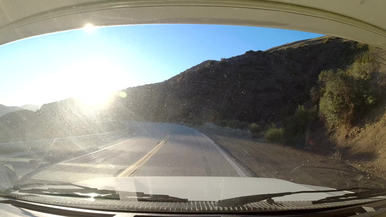 A windshield view in Coast California shows the road bending right around a hillside, with dry brush along the shoulder, intense low sun, and no visible people.