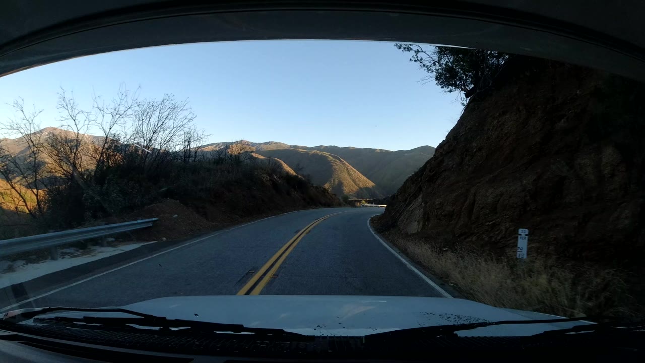 Forward windshield view of a Coast California two-lane road curving beside a rocky hillside and guardrail, with the vehicle hood at the bottom and no visible people.