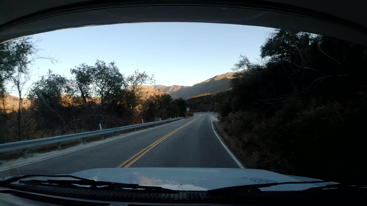 Forward vehicle view on a Coast California road with a guardrail at left, dark tree cover at right, sunlit hills in the distance, and no visible people.