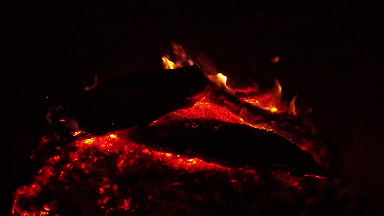 Close view of charred wood and red embers burning in Ojai, California, with small flames along the log edge and no visible people.