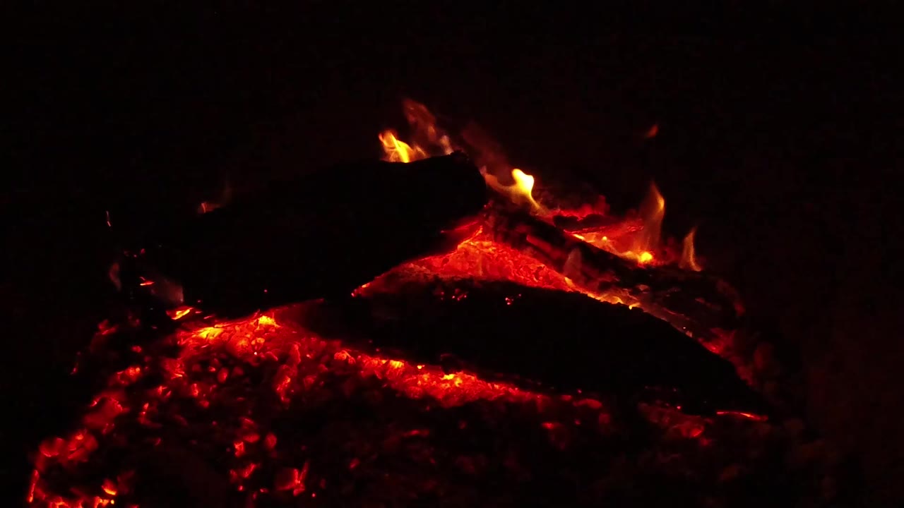Red ember bed glows beneath a blackened log in Ojai, California, with orange flame tips visible at the upper edge and no visible people.