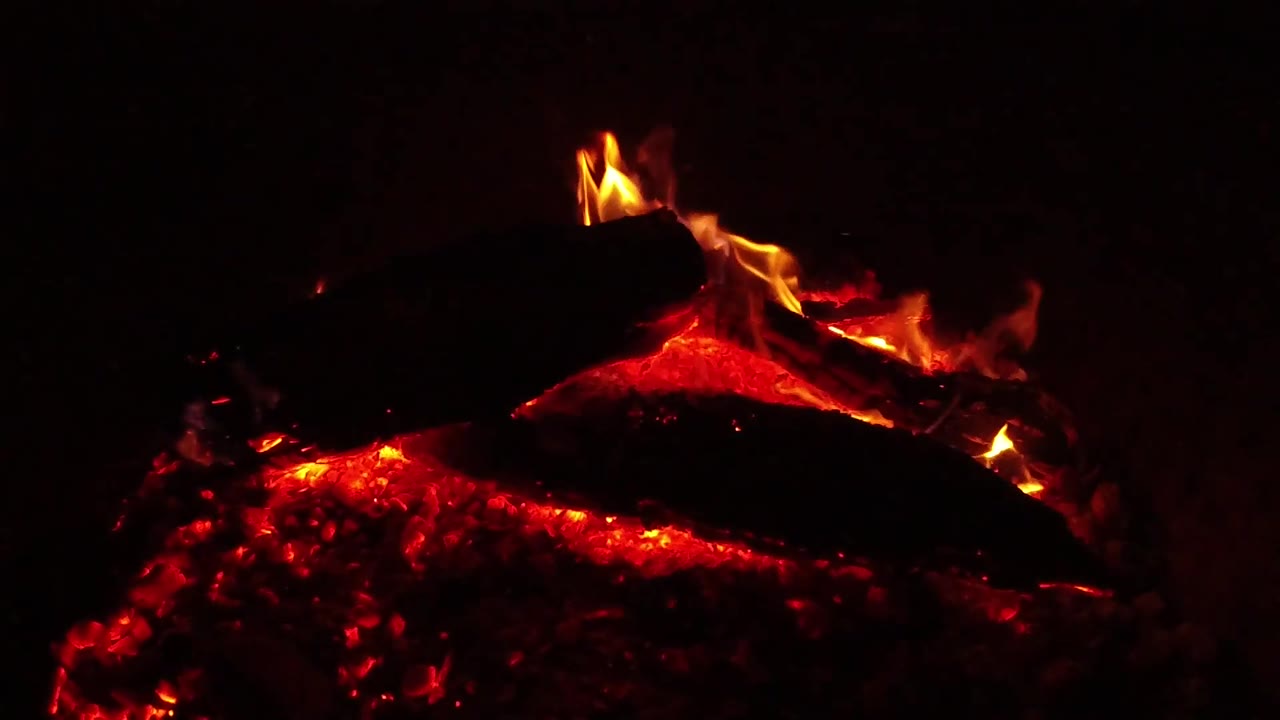 At 0:02, brighter yellow flames curl over the upper log while the lower embers glow red in the dark Ojai, California, bonfire scene, with no visible people.