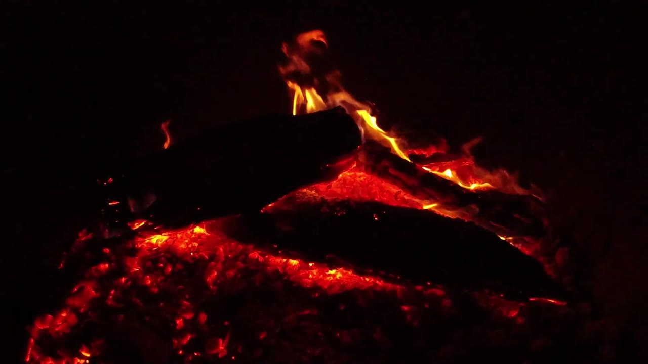 At 0:03, a dark log rests above a bright red ember bed while low orange flames appear near the top and right side, with no visible people in the Ojai, California frame.