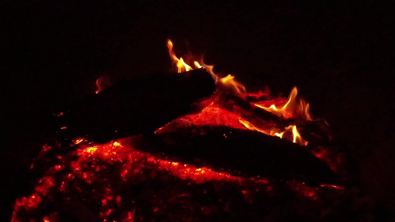 A close view of a campfire in Ojai, California, with red coals glowing under dark logs and small flames rising at the top, with no visible people.