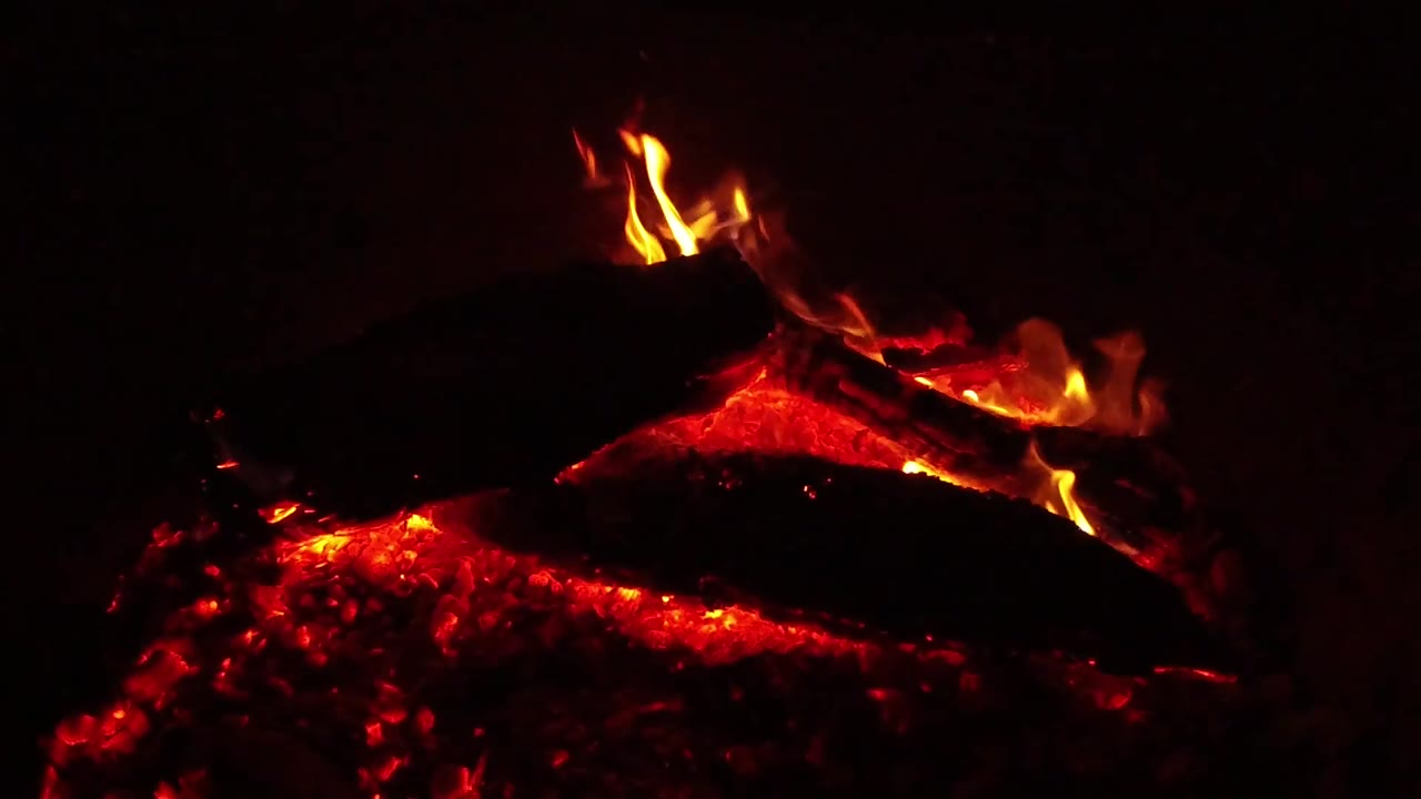 Bright orange flames lick around the right side of a blackened log over red coals in the Ojai night campfire, with the background falling into darkness and no visible people.