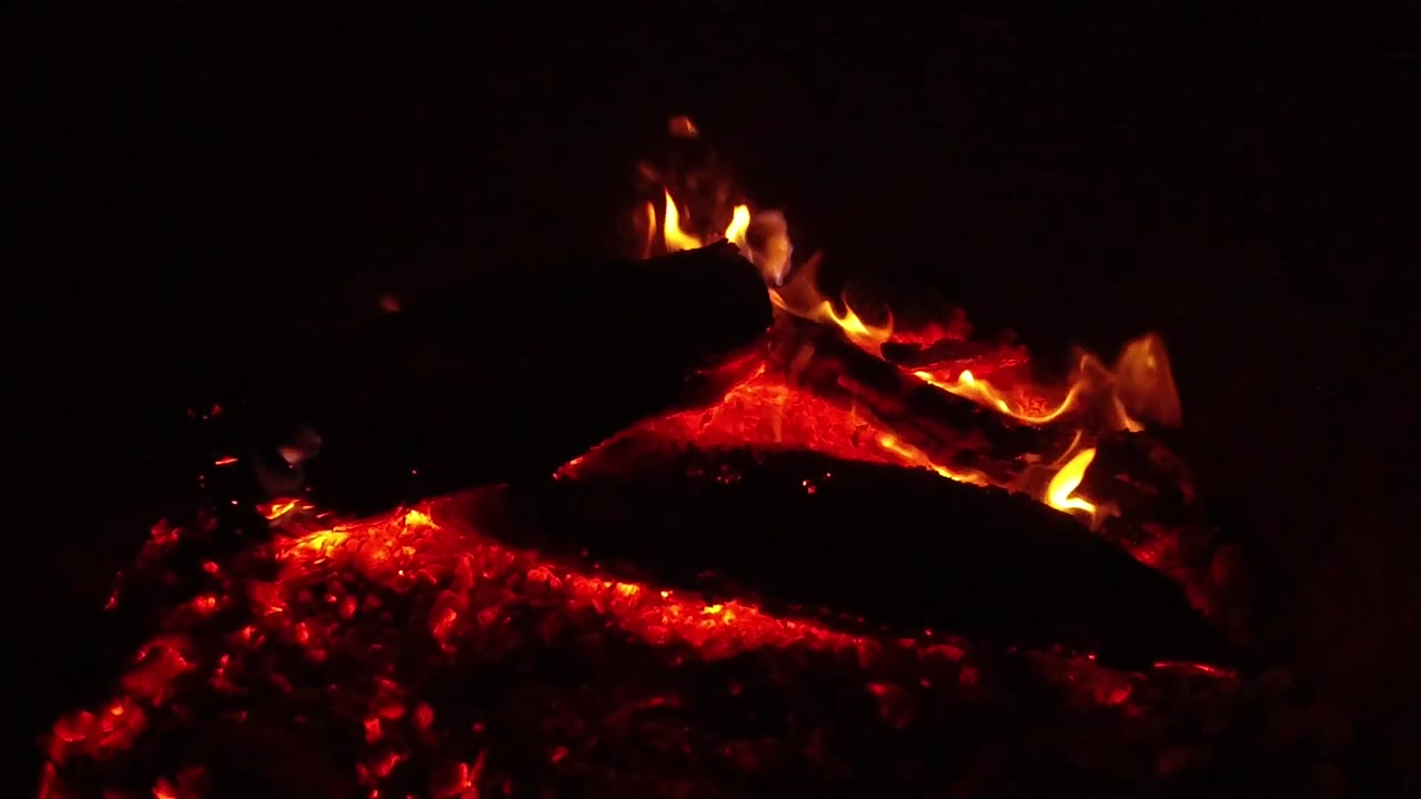 Low flames burn along the center and right edge of a dark log while red coals glow across the lower portion of the Ojai campfire frame, with no visible people.