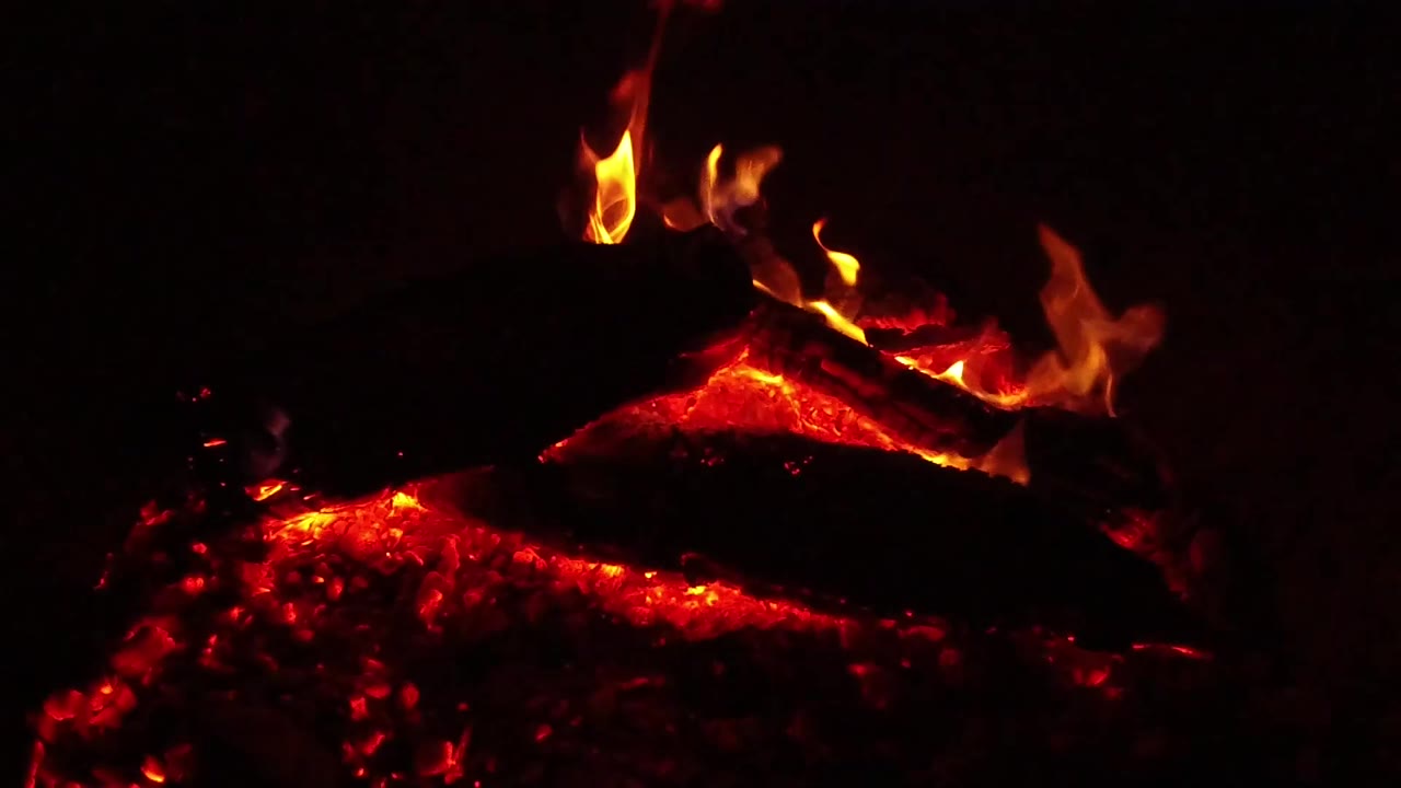 Orange flames stretch higher over a charred log while red coals glow below in the Ojai, California fire close-up, with no visible people.