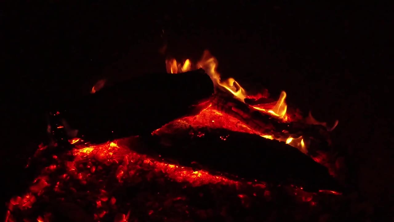 At 0:02, a dark log cuts across the fire bed as orange flame runs along its edge and embers glow underneath, with no visible people.