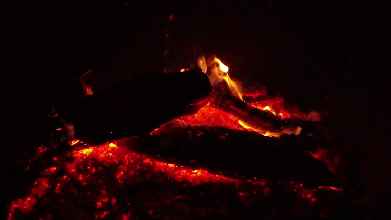 A close nighttime view in Ojai, California shows blackened logs resting over red-orange embers with small flames along the upper wood and no visible people.