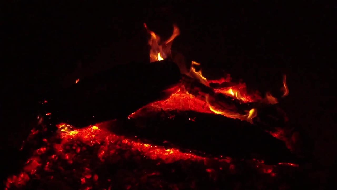 Small flames flare along the top of a blackened log above glowing coals in the Ojai night campfire, with no visible people.