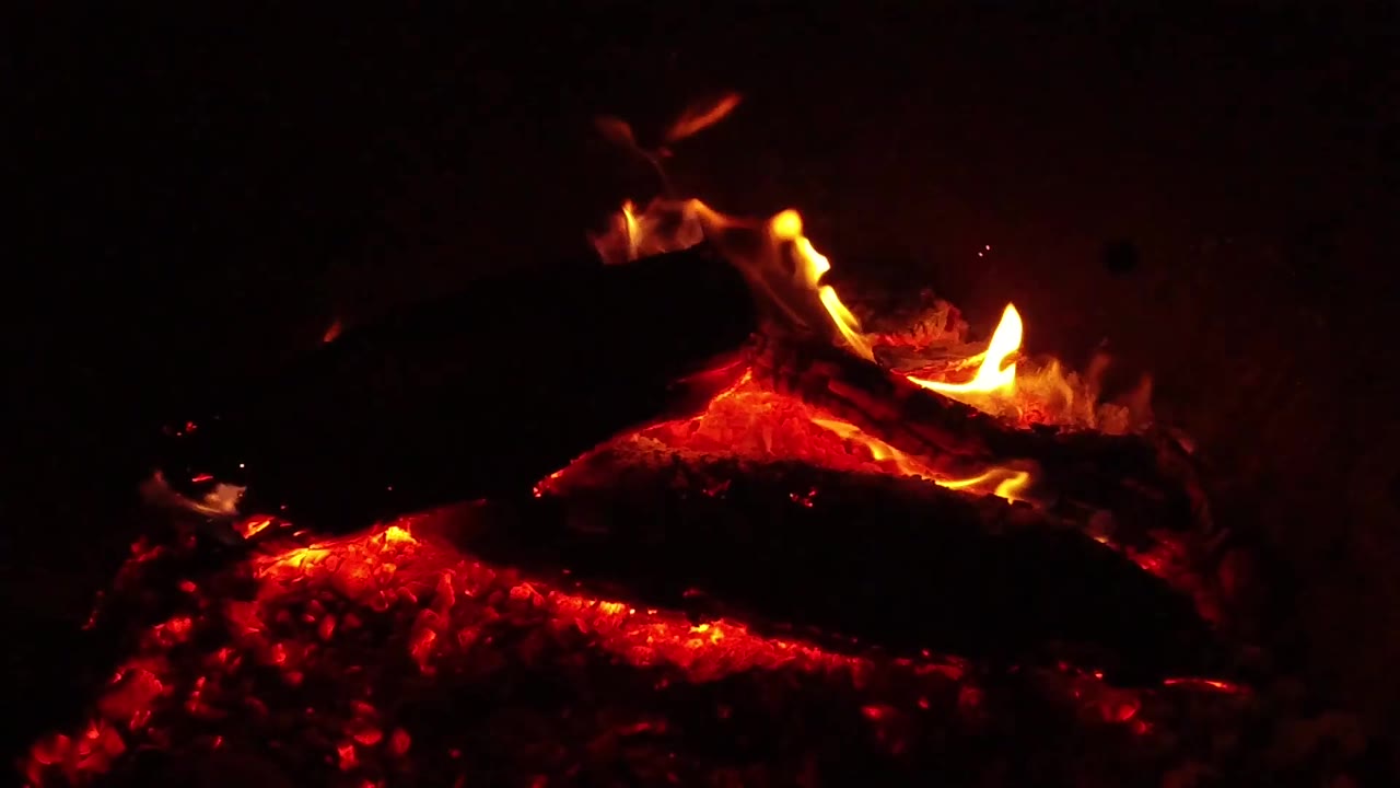 A brighter orange flame curls around the left side of the campfire log in Ojai, while red embers glow below and no people are visible.
