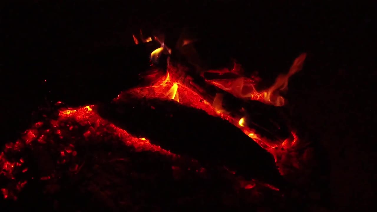 At 0:02, the coal bed glows red beneath a dark diagonal log while orange flame curls along the top in Ojai, California, with no visible people.
