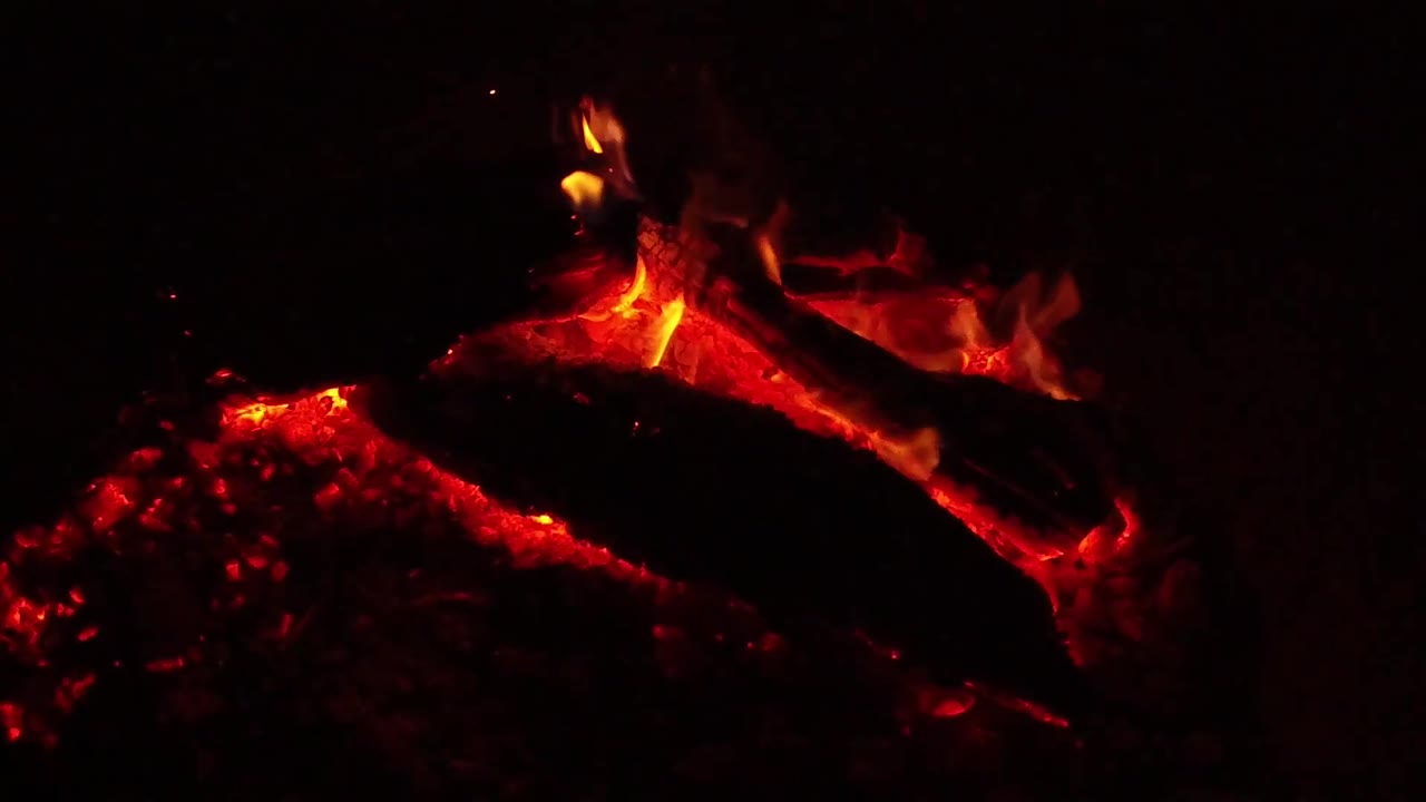 At 0:02, a taller flame lifts from the upper edge of the charred log above red embers in the Ojai night scene, with no visible people.