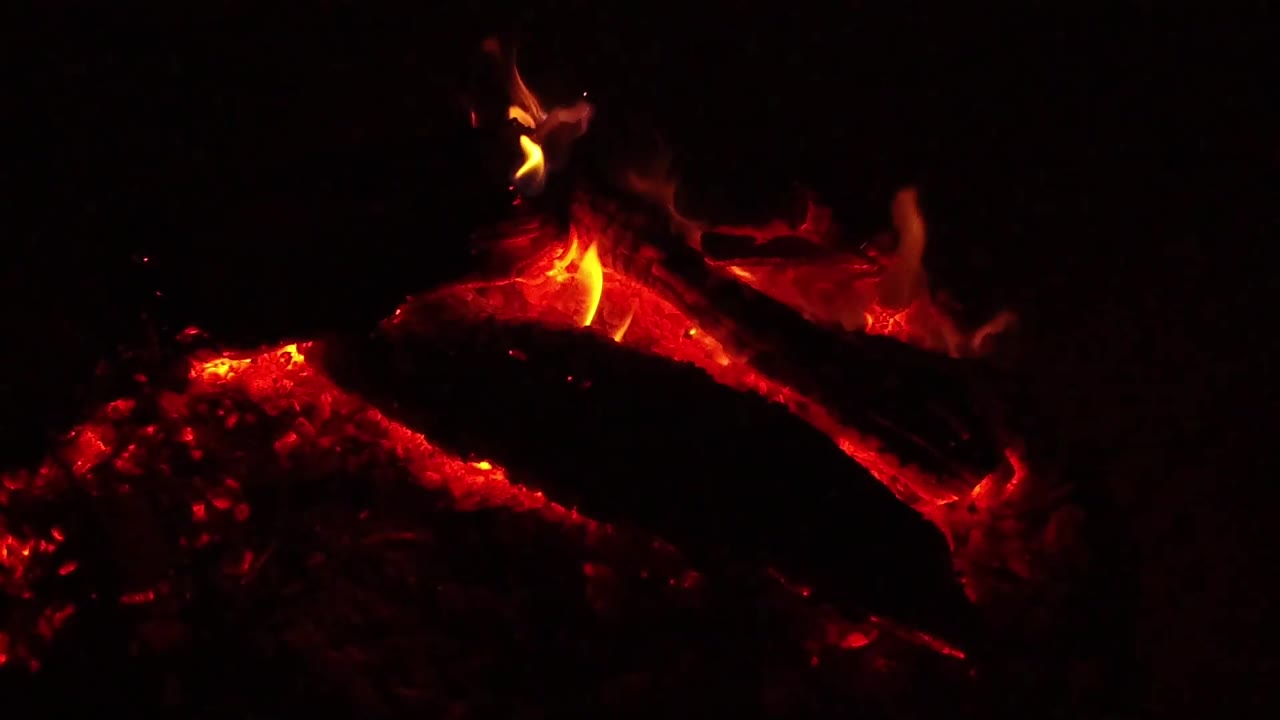 At 0:02 in Ojai, California, the blackened log spans the frame over red-orange embers, with a narrow flame tracing the upper surface and no visible people.