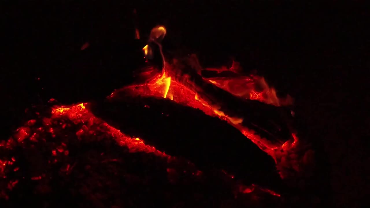 At 0:01, the Ojai, California fire holds on red coals and a thin yellow-orange flame curling above the central charred log, no visible people.