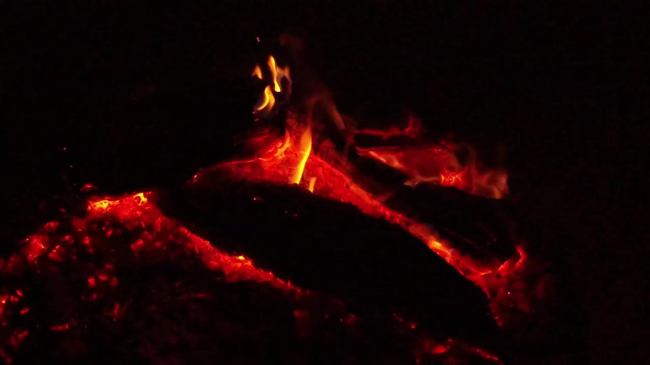 At 0:01, a charred log crosses the frame over red embers as a narrow flame lifts near the upper center in Ojai, California, with no visible people.