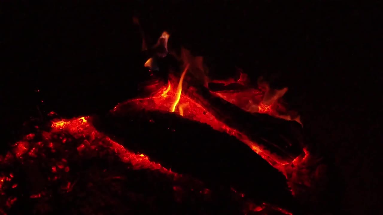 At 0:03, a vertical flame rises from the center of a glowing log while the surrounding frame stays nearly black in Ojai, California, with no visible people.
