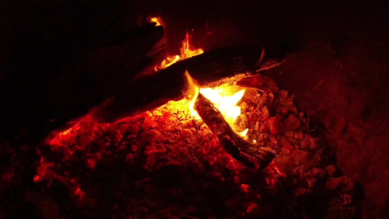 At 0:01, a brighter flame rises between two burned logs over red-orange embers in the Ojai nighttime campfire, with no visible people.