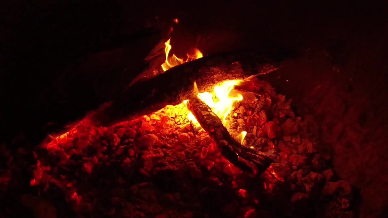 At 0:03, a smaller flame burns low under charred wood while red coals light the lower half of the Ojai campfire frame, with no visible people.