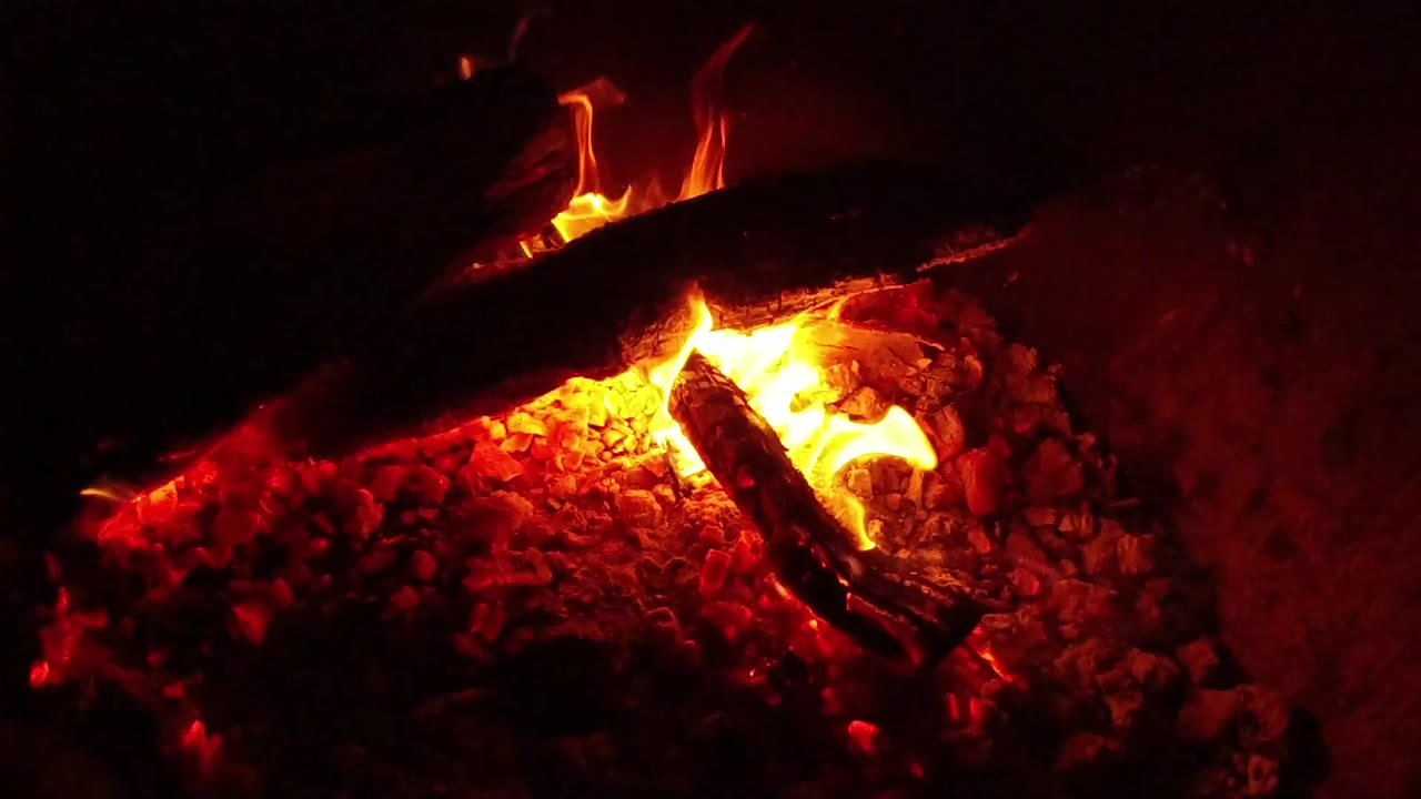 At 0:00, charred logs burn over a bed of red embers in Ojai, California, with orange flame rising near the center and no visible people.